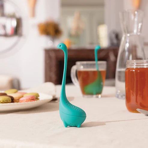 Teal dinosaur-shaped tea infuser on a table with tea and cookies.